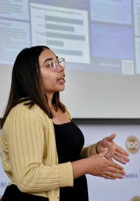 A young woman wearing a yellow sweater speaks and gestures